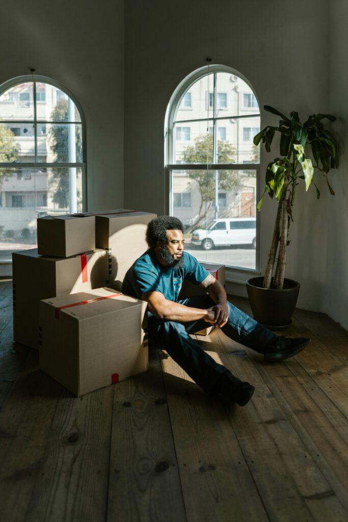 Reflective young man sitting among moving boxes in a sunlit room, contemplating new beginnings.